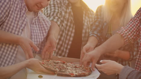 A-company-of-six-young-people-parses-pieces-of-hot-pizza-and-eats-together.-This-is-a-rooftop-party-with-a-beer.